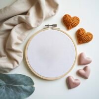 A beige cloth, yarn hearts, fabric hearts, and a green leaf surround a blank embroidery hoop—ideal for starting your own Custom Hand Embroidery Design (made to order) with complimentary draft service on a peaceful white surface.