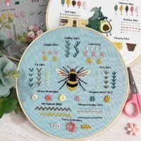 The Embroidery Stitch Practice Kit 3 features a round hoop with a central embroidered bee, surrounded by colourful flowers, leaves, and labelled examples of various stitch styles on light blue fabric.