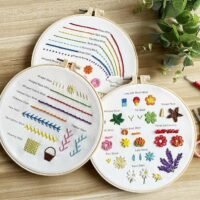 The Embroidery Stitch Practice Kit 4 features three hoops with colourful stitches and floral patterns on cream fabric, arranged on a wooden surface alongside green leaves and red scissors.