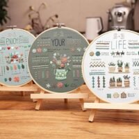 The Embroidery Stitch Practice Kit 1 features three wooden stand hoops with various stitched patterns, floral designs, and decorative text, beautifully displayed on a table with blurred kitchen items in the background.