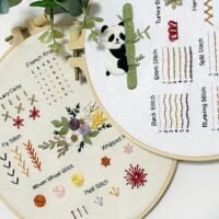 The Embroidery Stitch Practice Kit 6 includes two hoops: one with hand embroidery stitches and a panda with bamboo, and another featuring labelled stitches, a daisy, and a floral arrangement with colourful thread samples.
