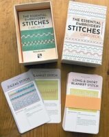 The Essential Embroidery Stitches Card Deck by Betty Barnden is shown on a wooden surface, featuring the box, an open box displaying cards, and three stitch instruction cards spread out.