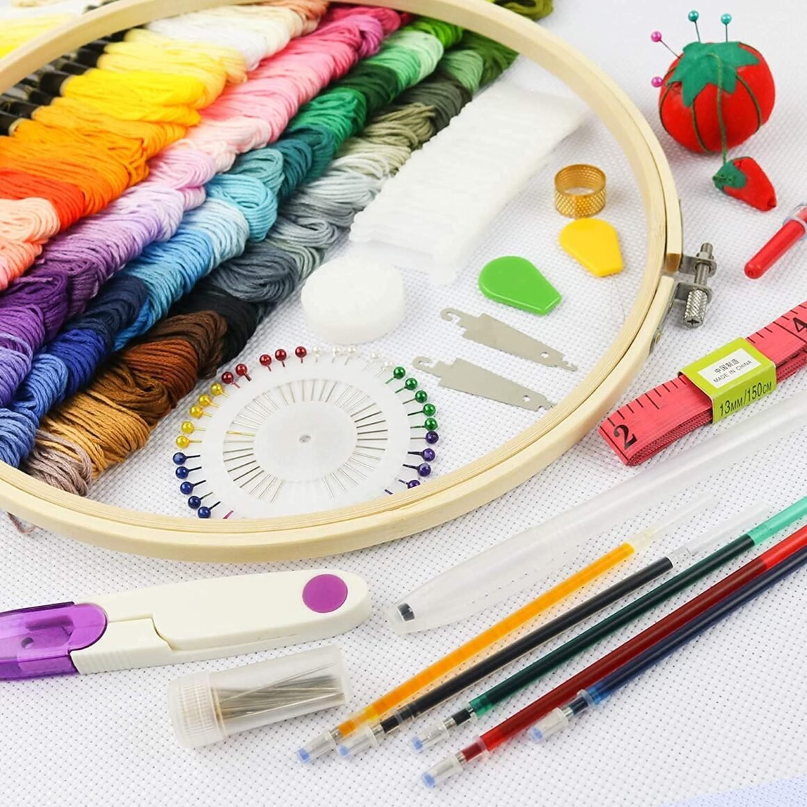 The Hand Embroidery Starter Kit (215 pieces) includes colourful threads, a wooden hoop, sewing tools, needles, pins, measuring tape, coloured pens, and a tomato-shaped pincushion arranged neatly on a white surface.