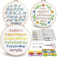 The Embroidery Stitch Practice Kit 10 features three hoops with stitched alphabets in assorted styles and colours, a floral wreath design, embroidery threads, needles, scissors, and an open instruction booklet.