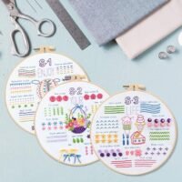 The Embroidery Stitch Practice Kit 13 features three hoops displaying vibrant embroidered samples—flowers, desserts, and patterns—on a light blue surface, with scissors, ruler, fabric, and thread spools arranged nearby.