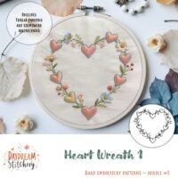 An embroidery hoop displays a heart-shaped floral wreath of pink hearts and flowers on white fabric. The text reads “Heart Wreath 1: Hand embroidery pattern.” Dried flowers and a pink cloth are nearby.
